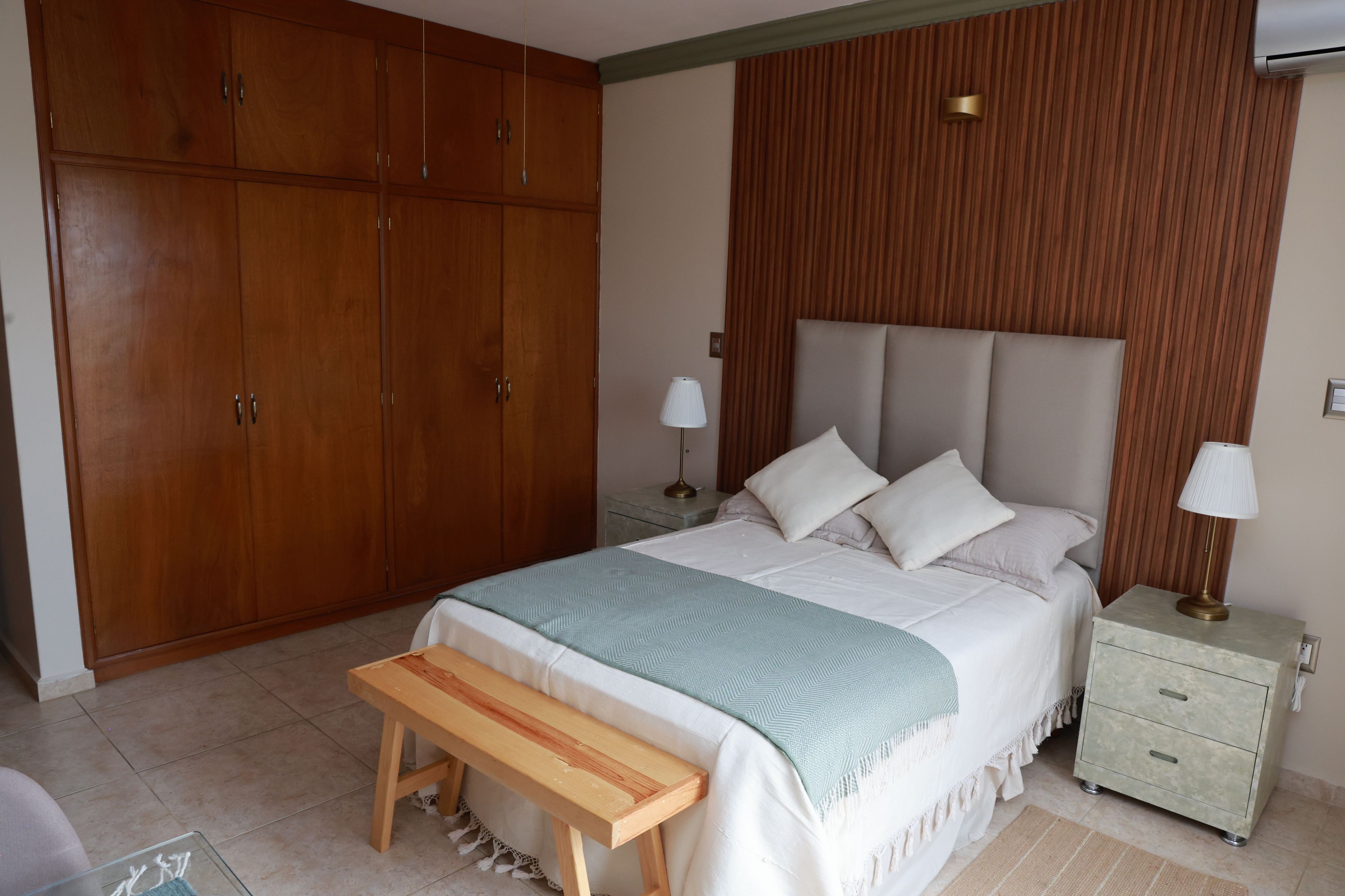 Un dormitorio con un amplio armario de madera, una cama con ropa de cama blanca, un banco y lámparas de noche.
