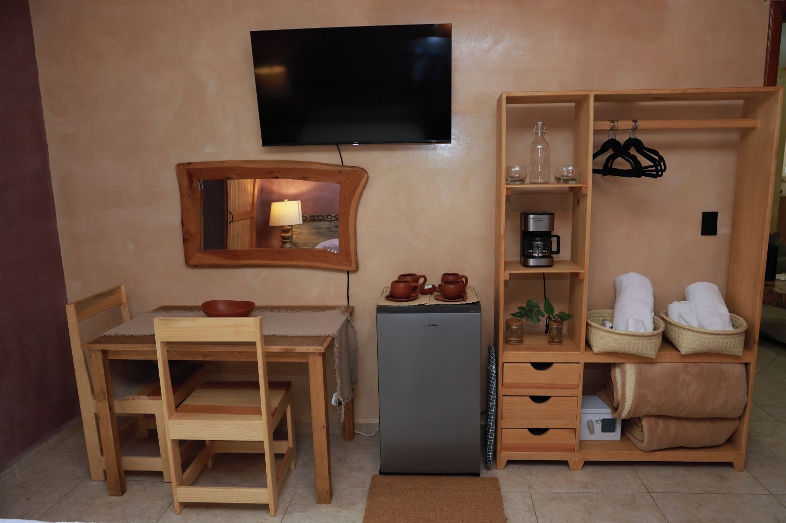 Habitación compacta con una mesa de madera, sillas, un refrigerador pequeño, un televisor montado en la pared y un estante con toallas y una cafetera.