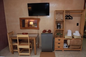 Habitación compacta con una mesa de madera, sillas, un refrigerador pequeño, un televisor montado en la pared y un estante con toallas y una cafetera.
