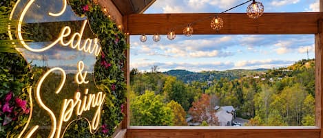 A wooden balcony with a neon sign reading 'Cedar & Spring' and a view of a forested landscape.