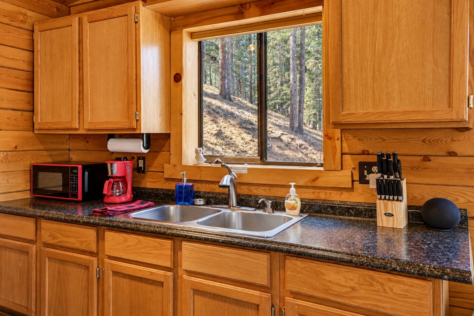 A wooden cabin kitchen with a window, a sink, a microwave, a coffee maker, and a knife block.