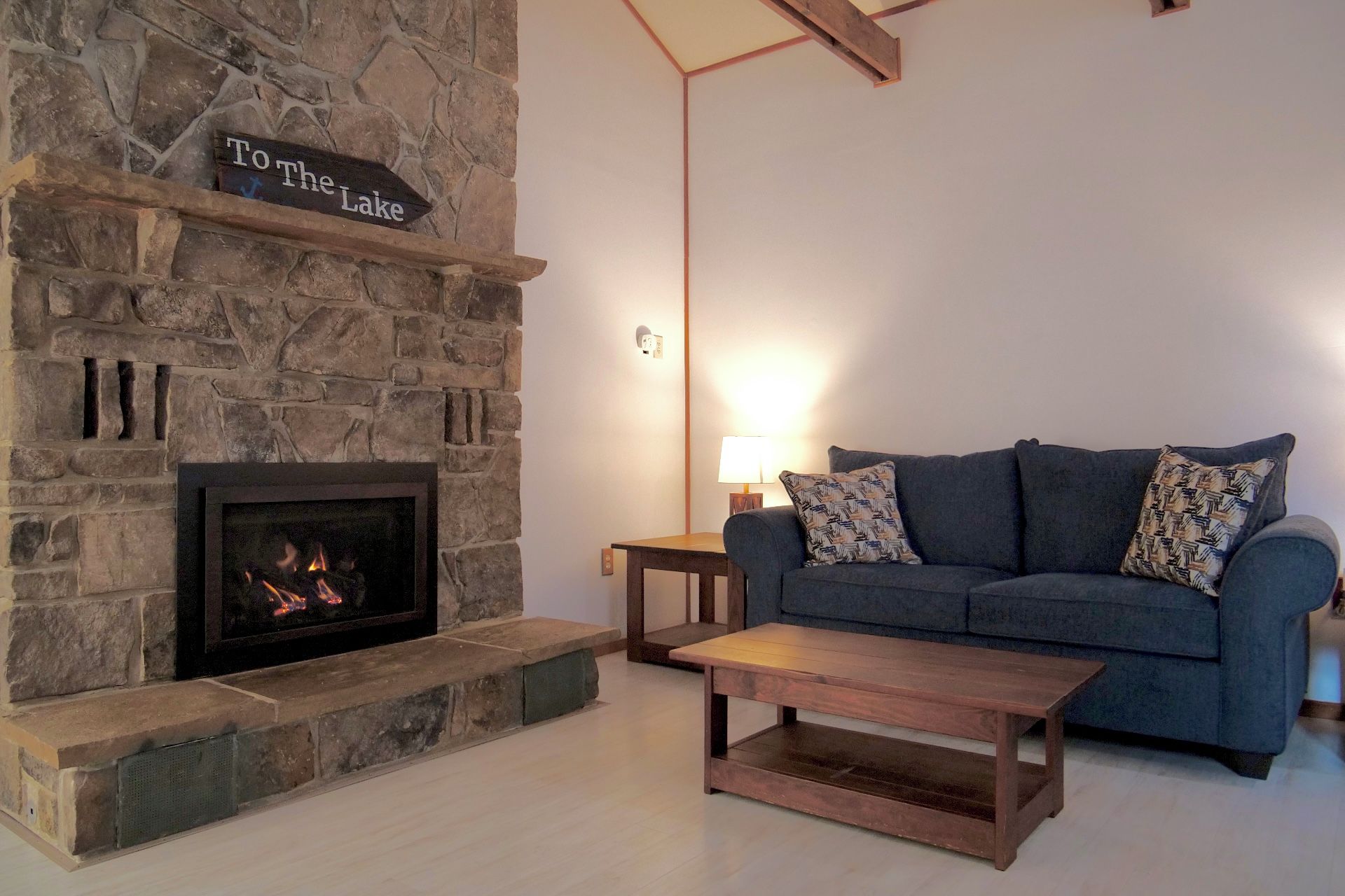 A stone fireplace with a sign reading 'To the Lake,' a blue sofa with patterned cushions, and a wooden coffee table.