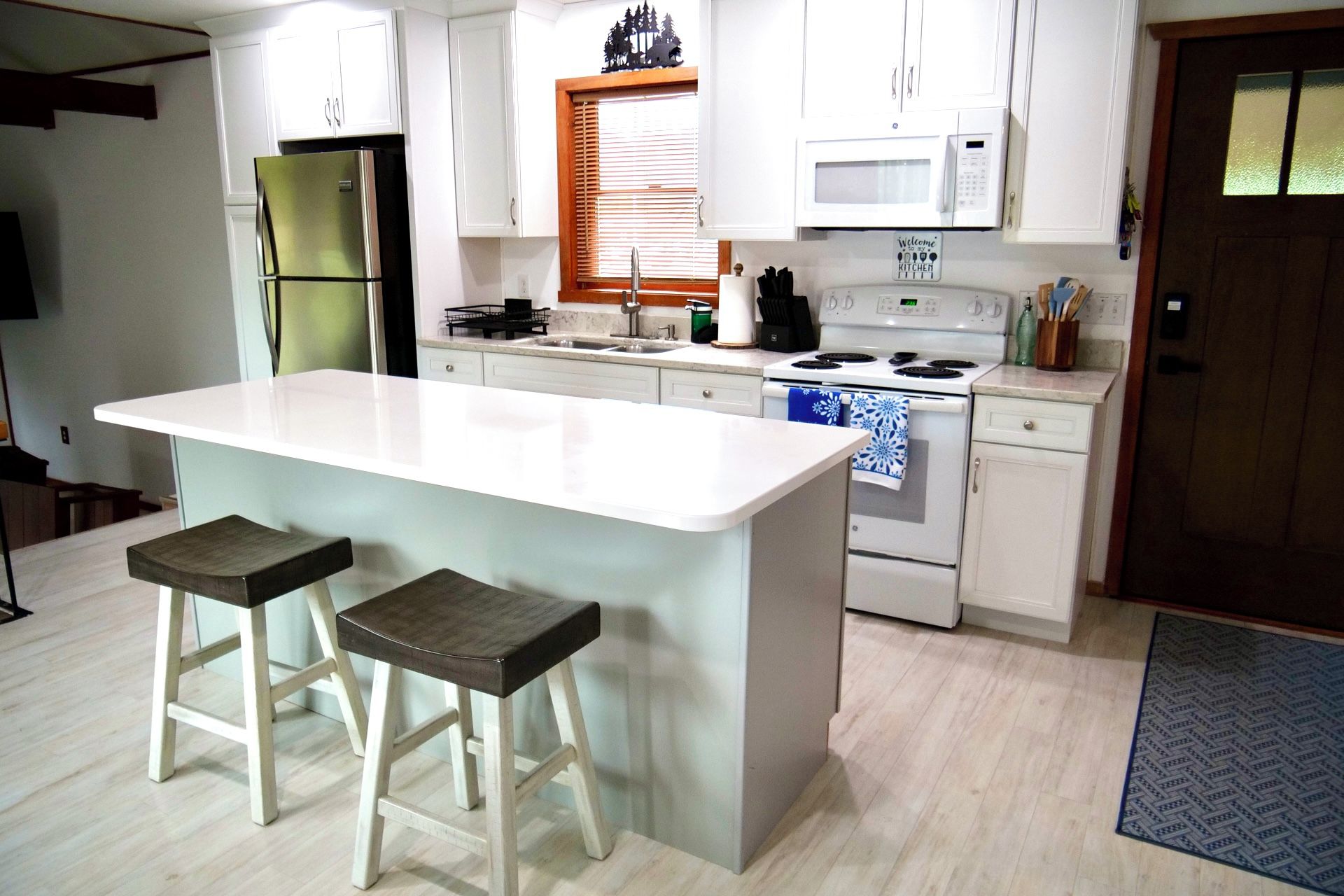 A kitchen with a central island, two bar stools, a refrigerator, a microwave, a stove, and a window with blinds.
