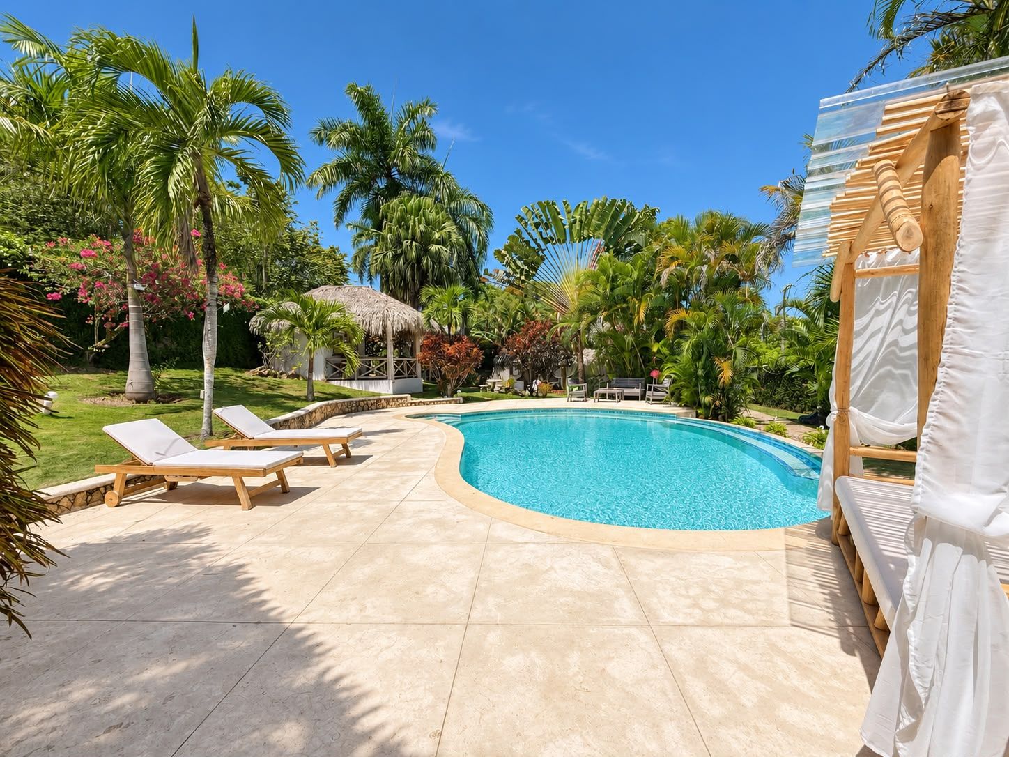 A swimming pool with lounge chairs, surrounded by palm trees and a thatched-roof structure.