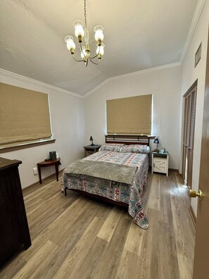 Quarto com cama, mesas de cabeceira, escrivaninha, cadeira, luminária de mesa, relógio e lustre.