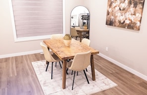 Een eetgedeelte met een houten tafel, vier stoelen, een vaas en een schilderij aan de muur.
