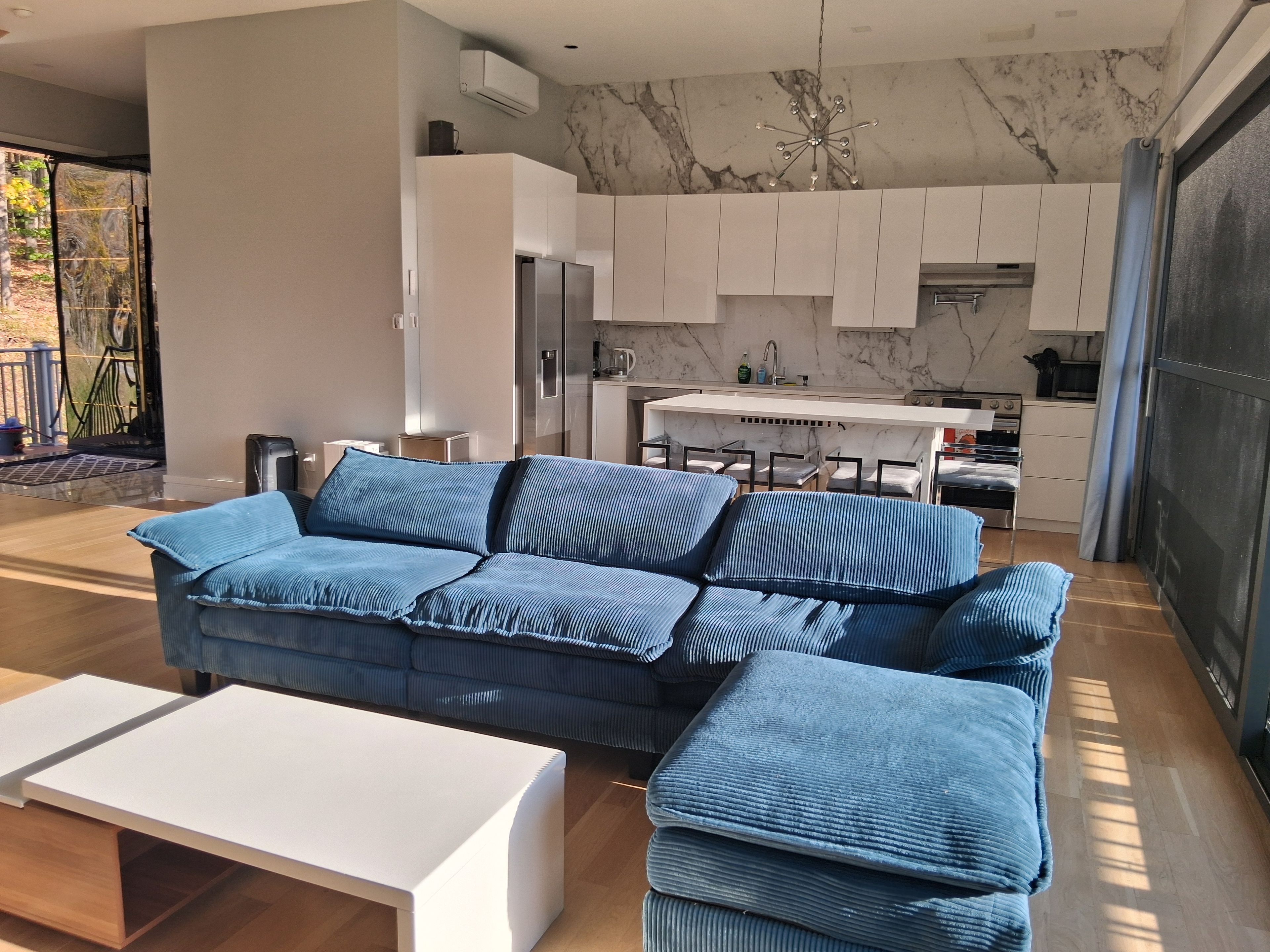 A modern living room with a sectional sofa, a white coffee table, and a kitchen area with white cabinets and a marble backsplash.