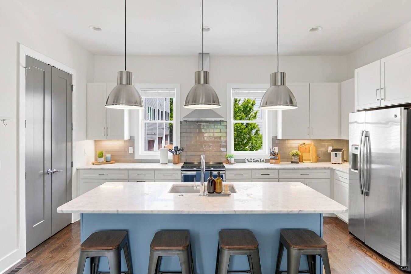 Une cuisine moderne dotée d’un îlot central, d’appareils électroménagers en acier inoxydable, de luminaires suspendus et offrant une vue sur la verdure environnante.