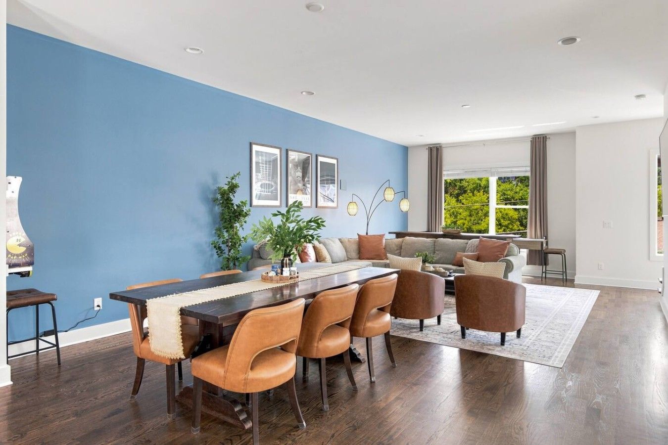 A modern living room with a dining area, featuring a blue accent wall, wooden flooring, and a large window providing a view of greenery.