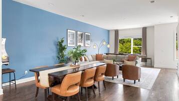 A modern living room with a dining area, featuring a blue accent wall, wooden flooring, and a large window providing a view of greenery.