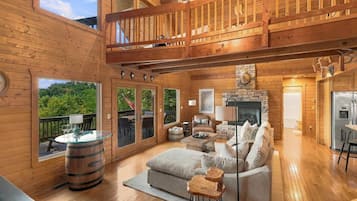 Een ruim houten interieur met een open haard, zithoek en een balkon met uitzicht op groen.