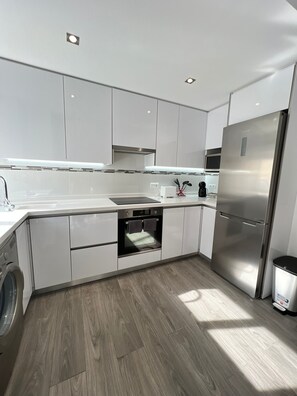 A modern kitchen with white cabinets, a stainless steel refrigerator, a built-in oven, and a washing machine.