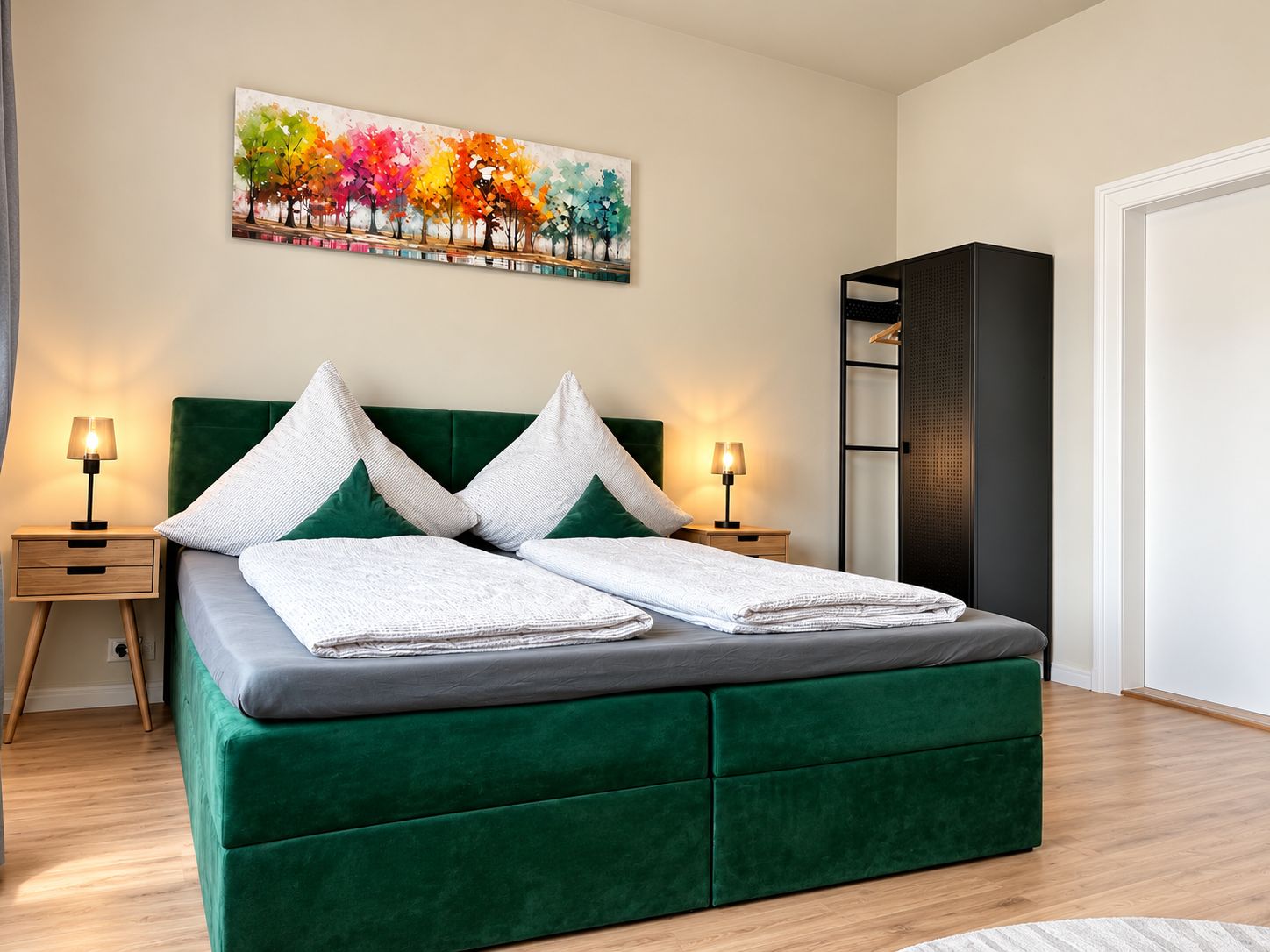 A modern bedroom with a green upholstered bed, two bedside tables with lamps, a large abstract painting on the wall, and a black wardrobe.