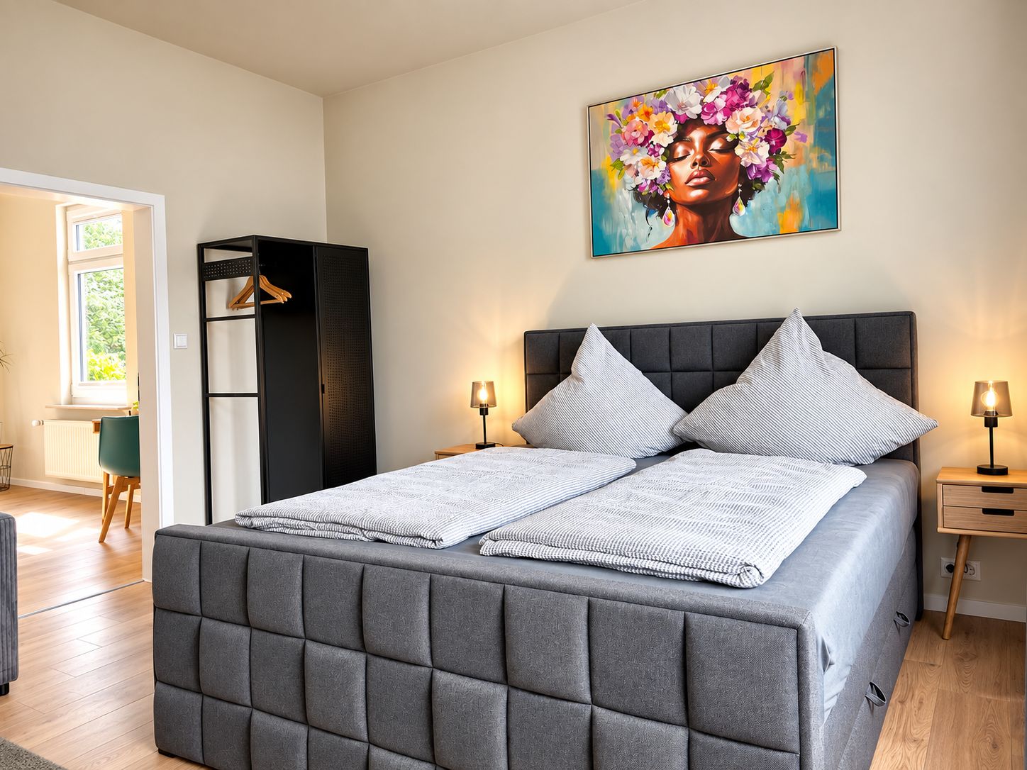 A modern bedroom with a grey upholstered bed, a large painting above the bed, and a wardrobe to the left.