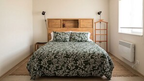 Een slaapkamer met een houten hoofdbord, een bed met een bloemenbedekking, een nachtkastje, een kapstok en een raam met jaloezieën.