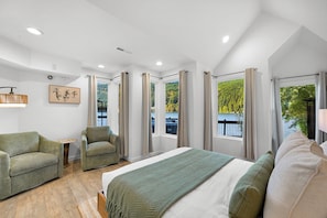 Un dormitorio moderno con una cama, dos sillas, una mesita y vistas a un lago y un bosque.