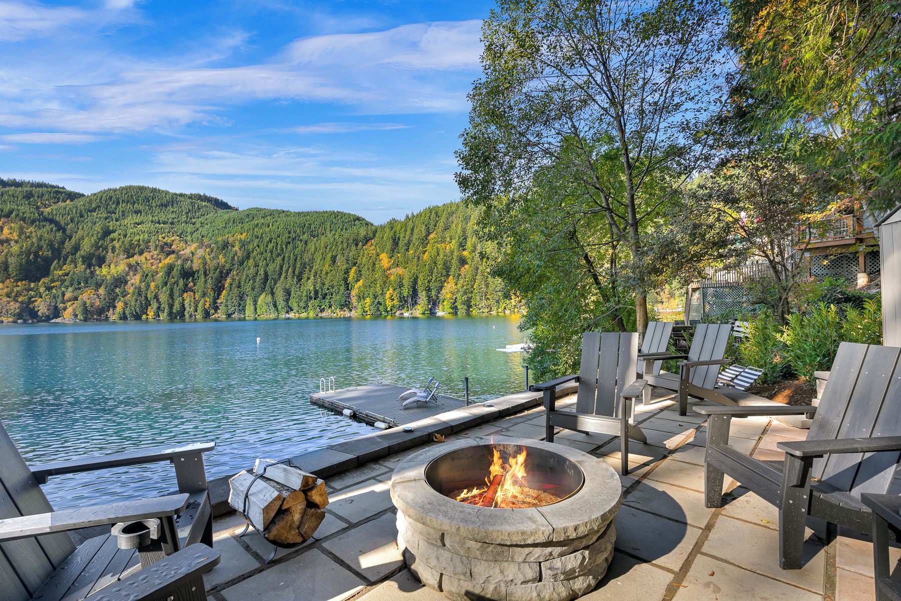 A lakeside deck with a fire pit, outdoor seating, and a view of the lake and forested hills.
