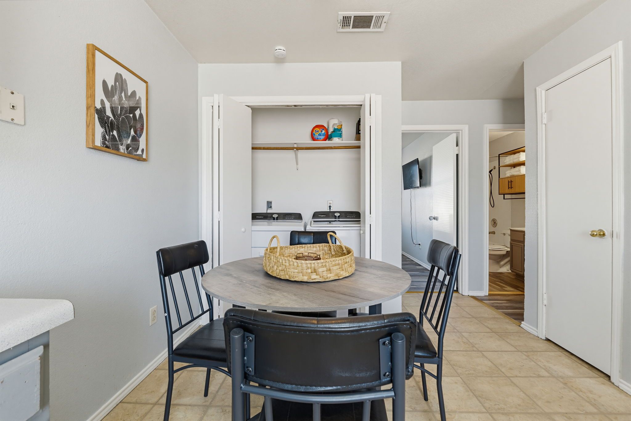 A dining area with a round table, four chairs, a woven basket, and a kitchen in the background.