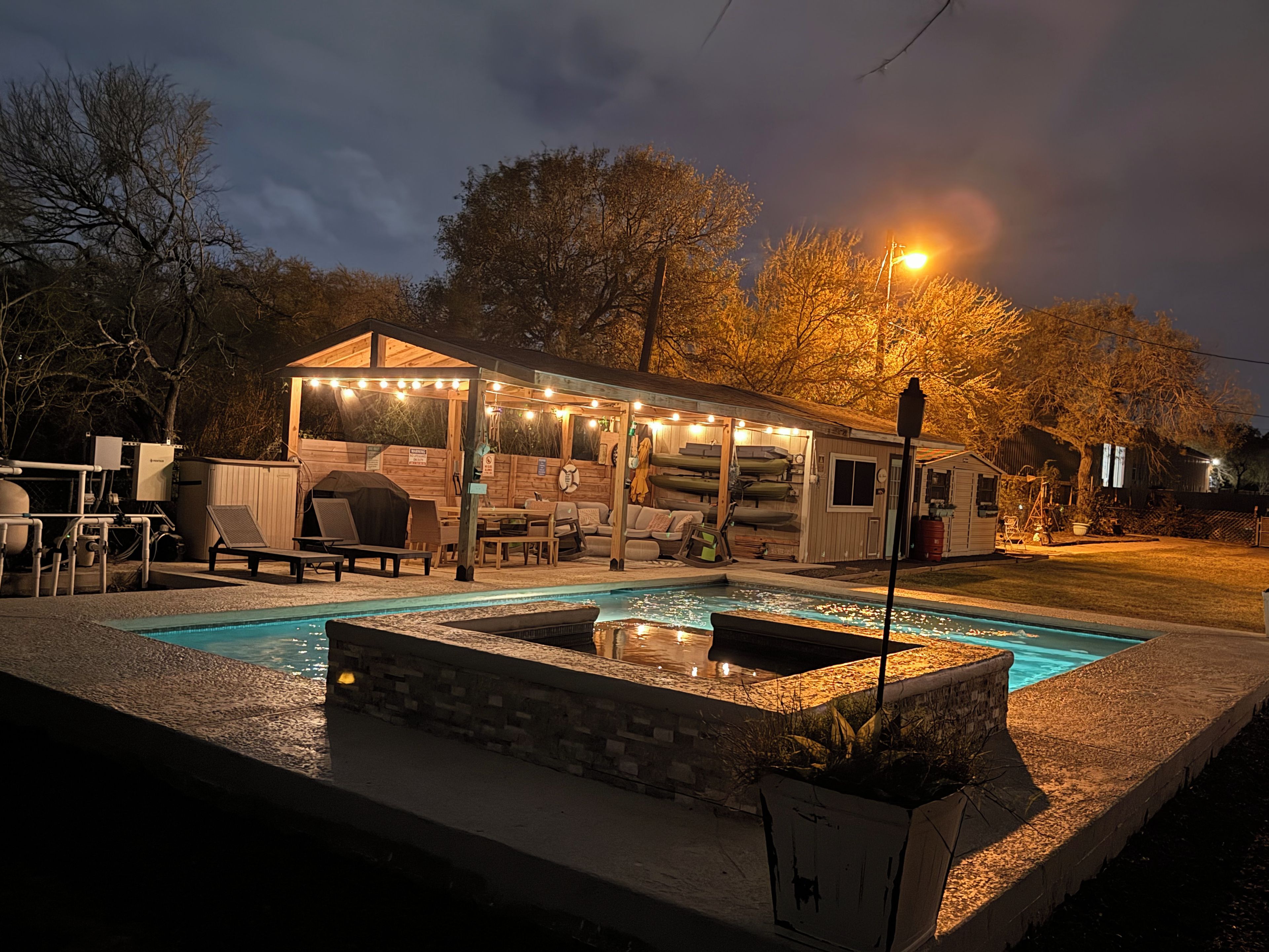Un área de piscina al aire libre con zona de estar techada, una parrilla de barbacoa y un cobertizo con kayaks.