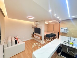 Ein modernes Hotelzimmer mit einer Couch, einem Fernseher, einem Bett, einer kleinen Küchenzeile und einem Essbereich.