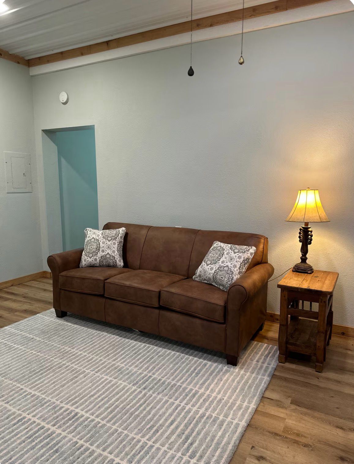 A brown sofa with patterned cushions, a small wooden side table with a lamp, and a white rug in a room with wooden beams on the ceiling and a light blue door.