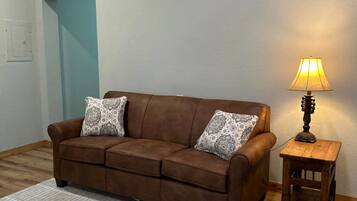 A brown sofa with patterned cushions, a small wooden side table with a lamp, and a white rug in a room with wooden beams on the ceiling and a light blue door.