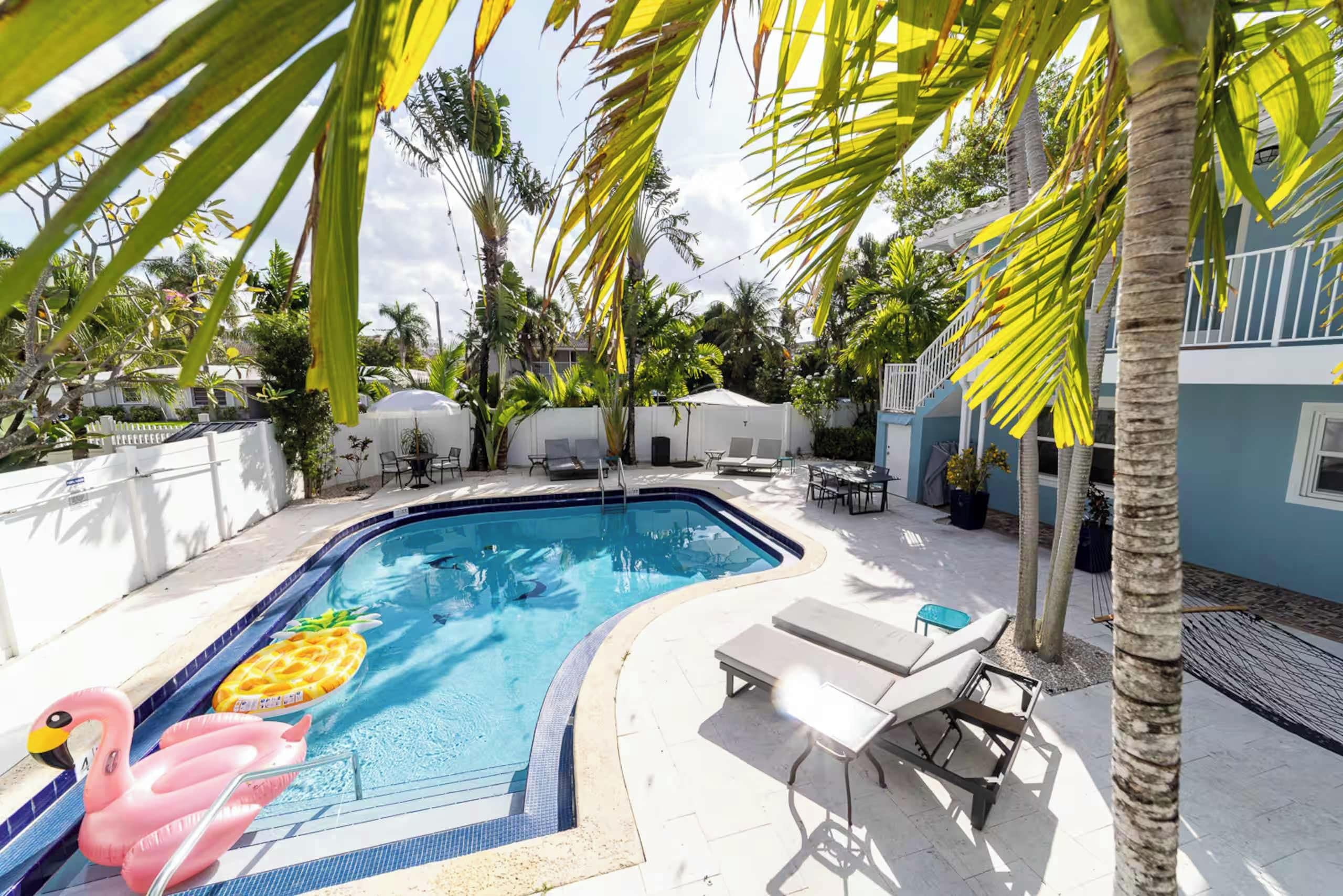 A swimming pool with lounge chairs and umbrellas, surrounded by palm trees and a white fence.