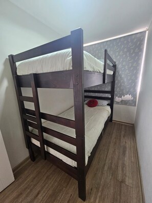 Una cama litera con estructura de madera y ropa de cama blanca, ubicada en una habitación con papel tapiz estampado y suelo de madera.