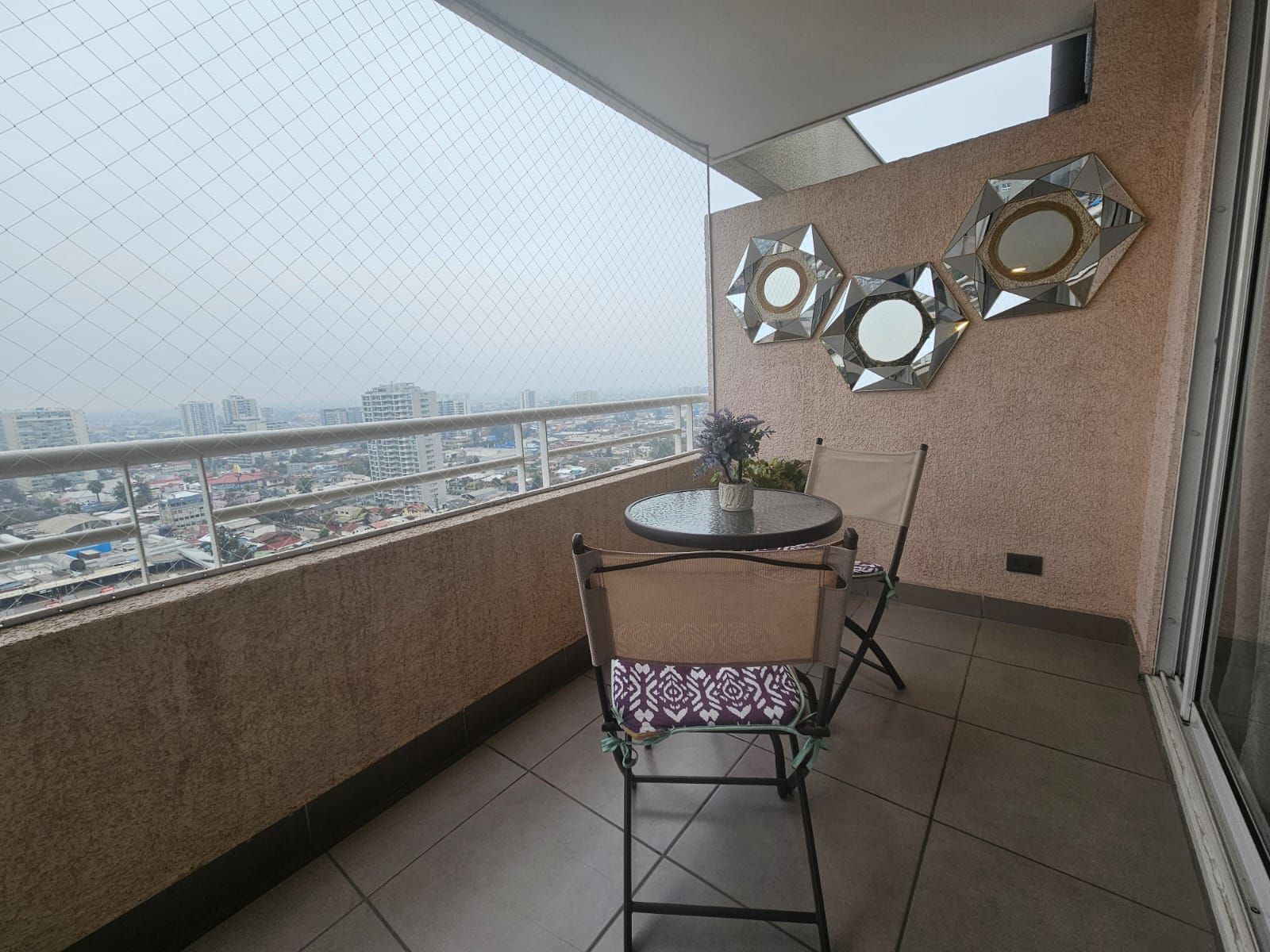 Un balcon avec une table ronde et deux chaises, un miroir décoratif fixé au mur et une vue sur le paysage urbain à travers une clôture en treillis métallique.