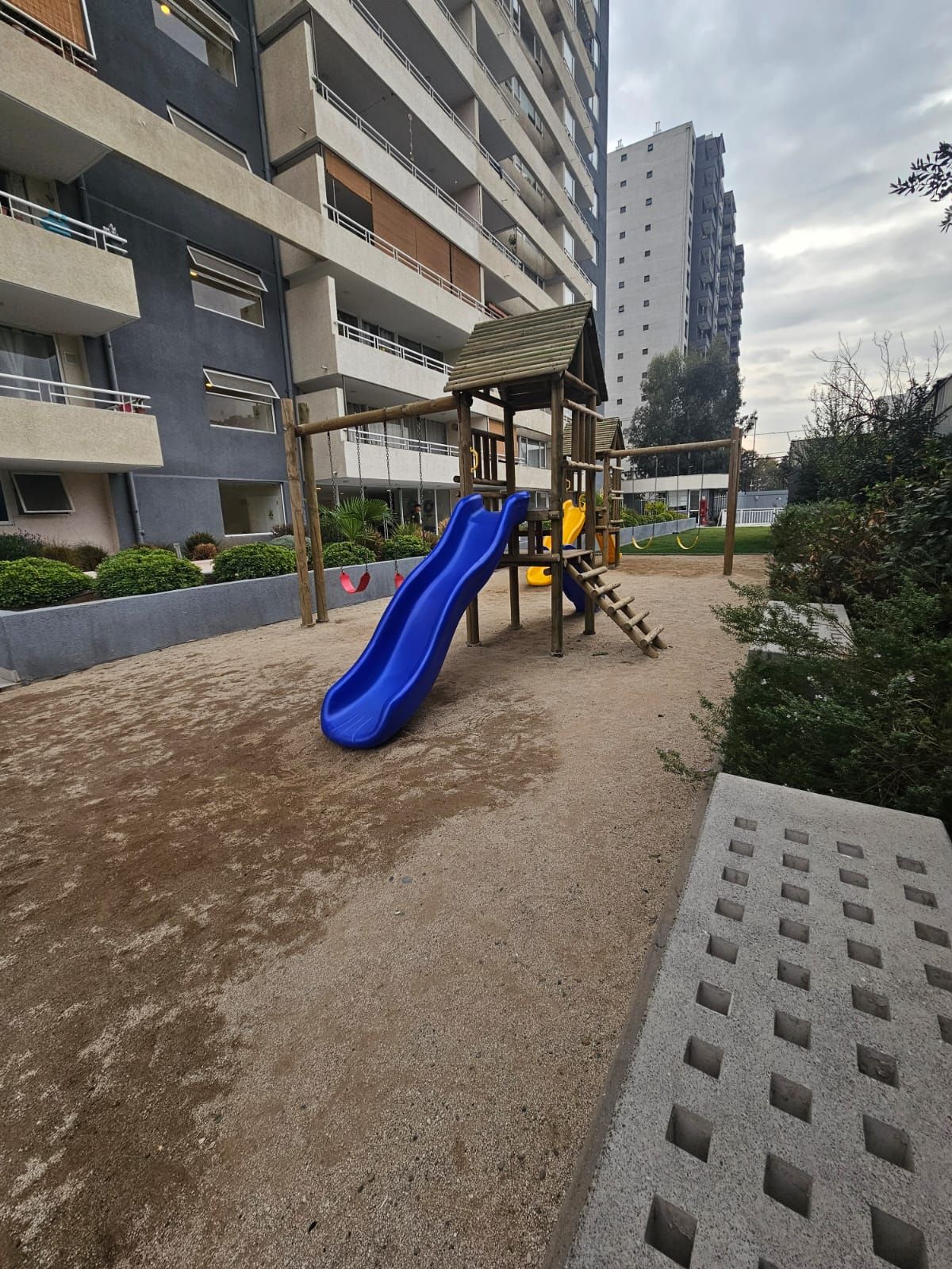 Leikkikentällä on sininen ja keltainen liukumäki, ja sen ympärillä on rakennus, jossa on useita parvekkeita.