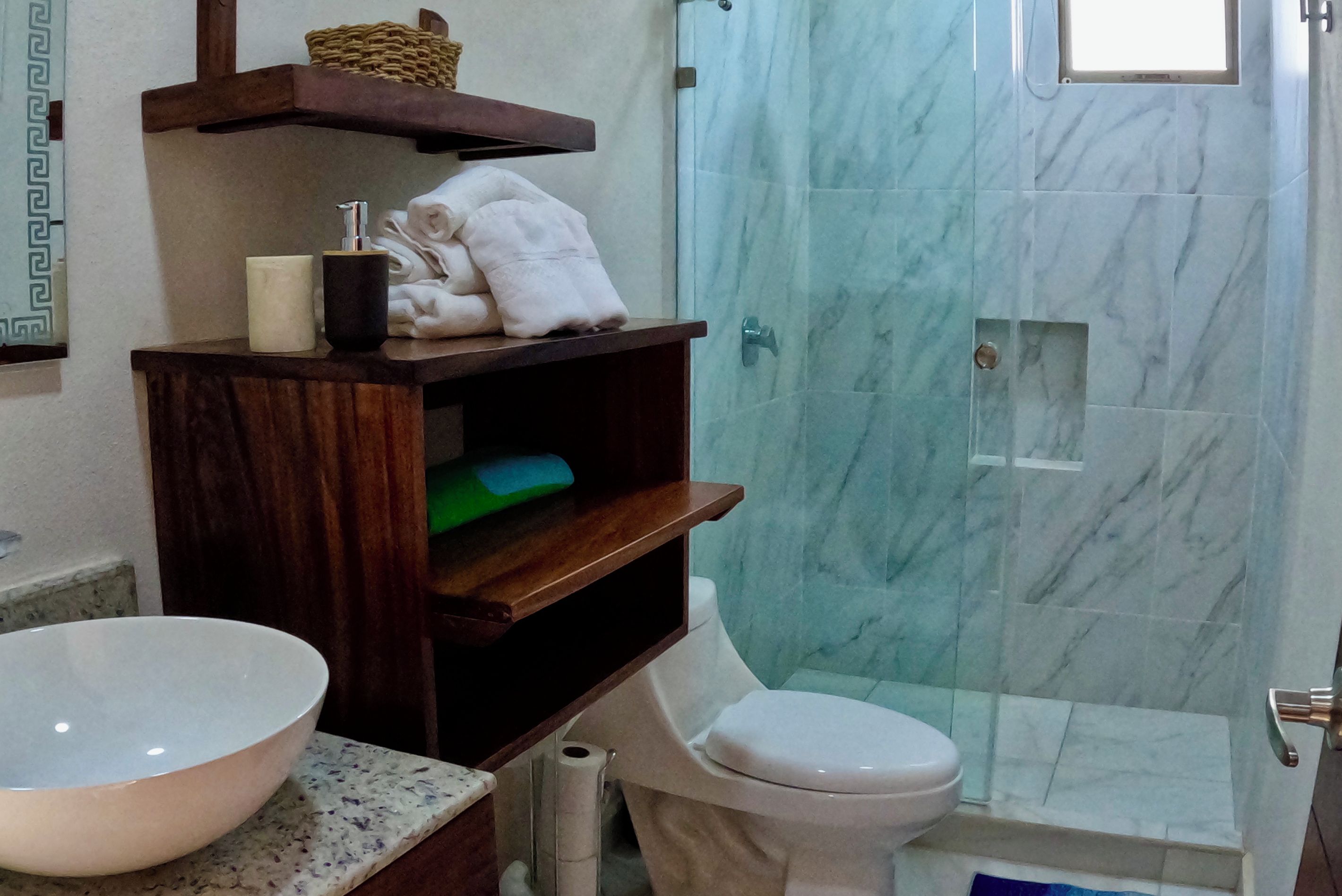 Un baño con ducha de mármol, inodoro blanco, tocador de madera con lavabo y estante con toallas y artículos de aseo.