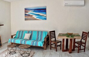 Una sala de estar con un sofá, una mesa de comedor con sillas, un cuadro en la pared y un equipo de aire acondicionado.