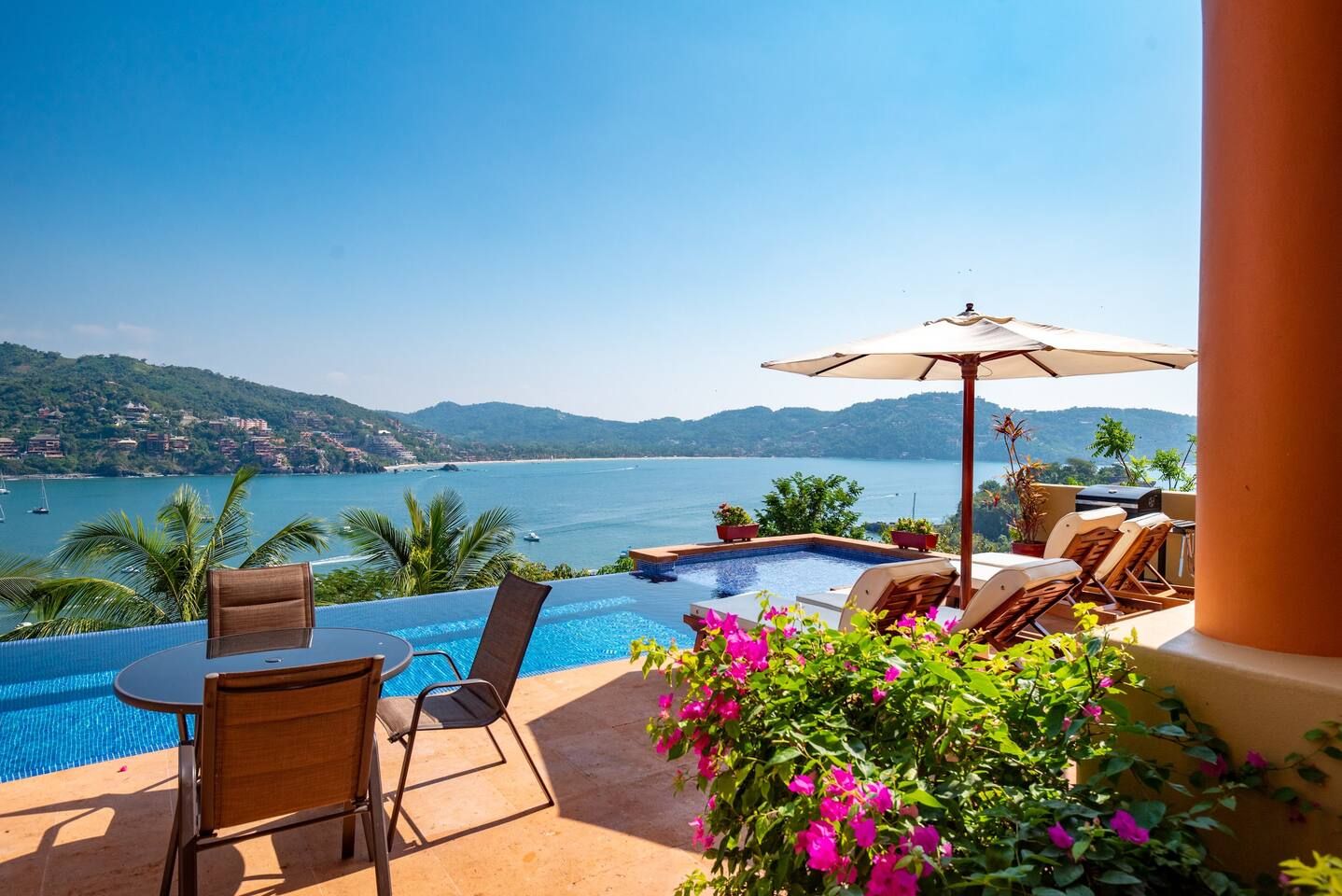 A poolside area with a view of the sea, a table, chairs, and a barbecue grill.