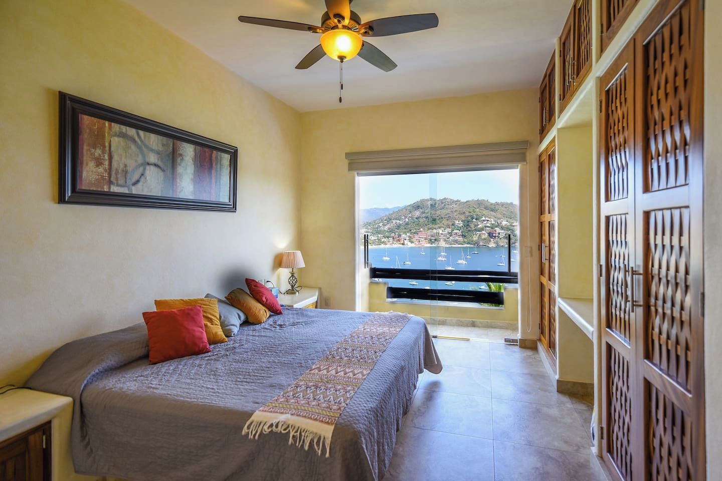 A bedroom with a bed, a nightstand, a lamp, a ceiling fan, and a view of the sea through a balcony.