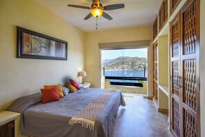 A bedroom with a bed, a nightstand, a lamp, a ceiling fan, and a view of the sea through a balcony.