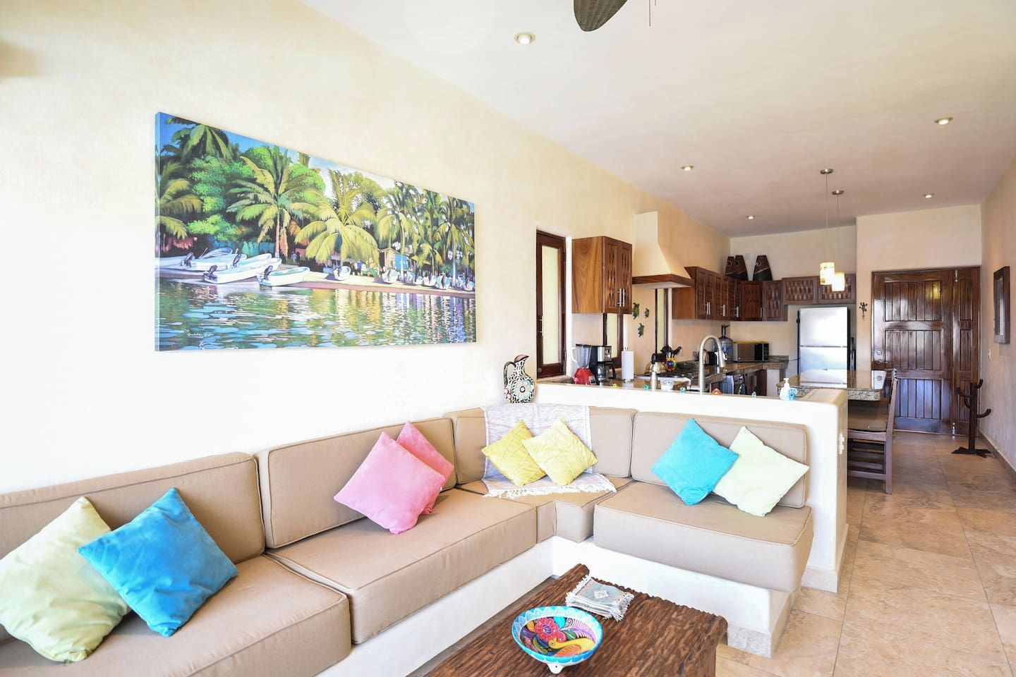 Una sala de estar con un sofá, cojines coloridos, una mesa de centro y un cuadro de una escena tropical.