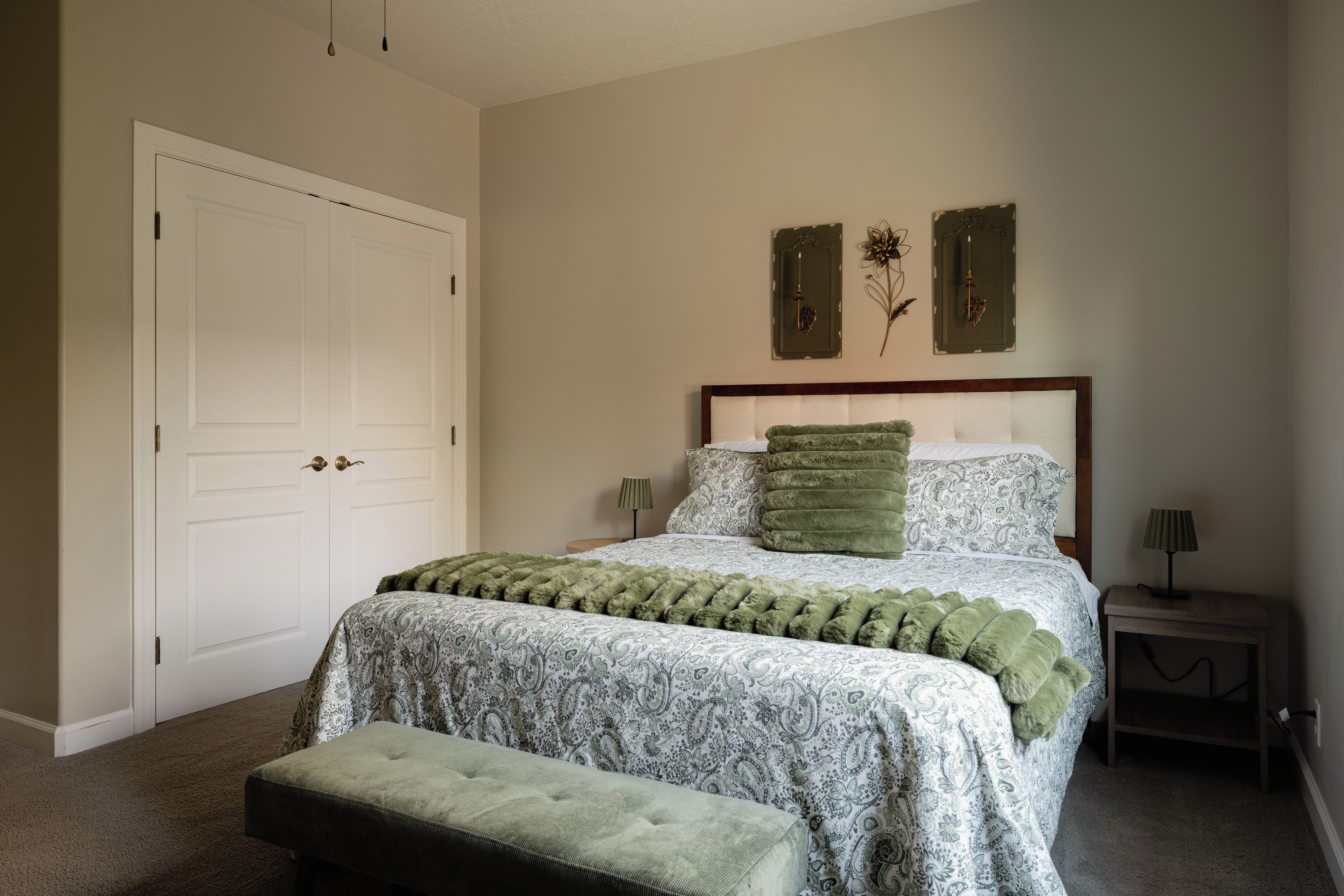 Une chambre avec un lit double, un plaid vert, un coussin vert assorti, une table de chevet avec une lampe et une porte dotée de poignées en laiton.