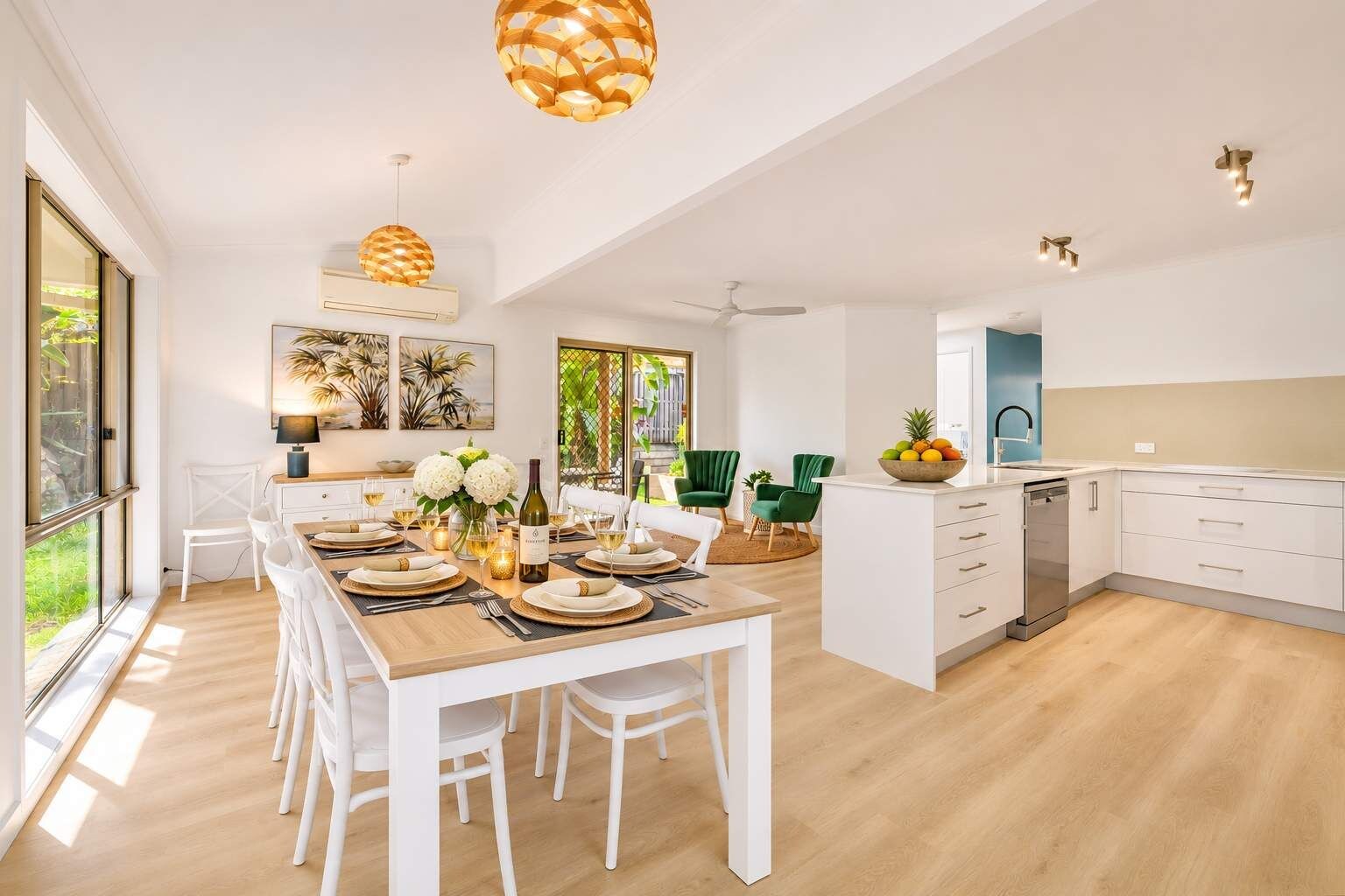 A modern kitchen with white cabinetry, a dining area with a wooden table set for six, and a view of the outdoors through large windows.