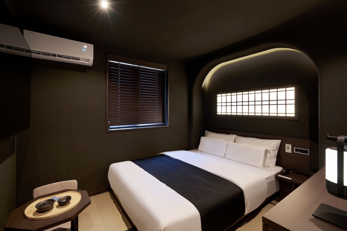 A hotel room with a bed, bedside table, lamp, air conditioning unit, and a window with blinds.