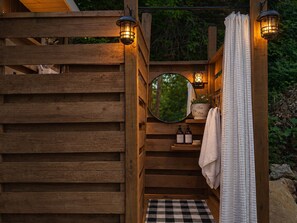 A wooden outdoor shower with a checkered floor, a mirror, a towel, and two bottles of shampoo.