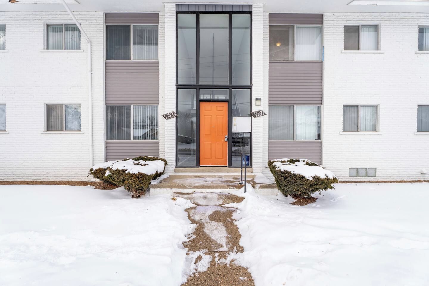 A multi-story building with a central entrance, a snow-covered pathway, and two snow-covered bushes on either side.