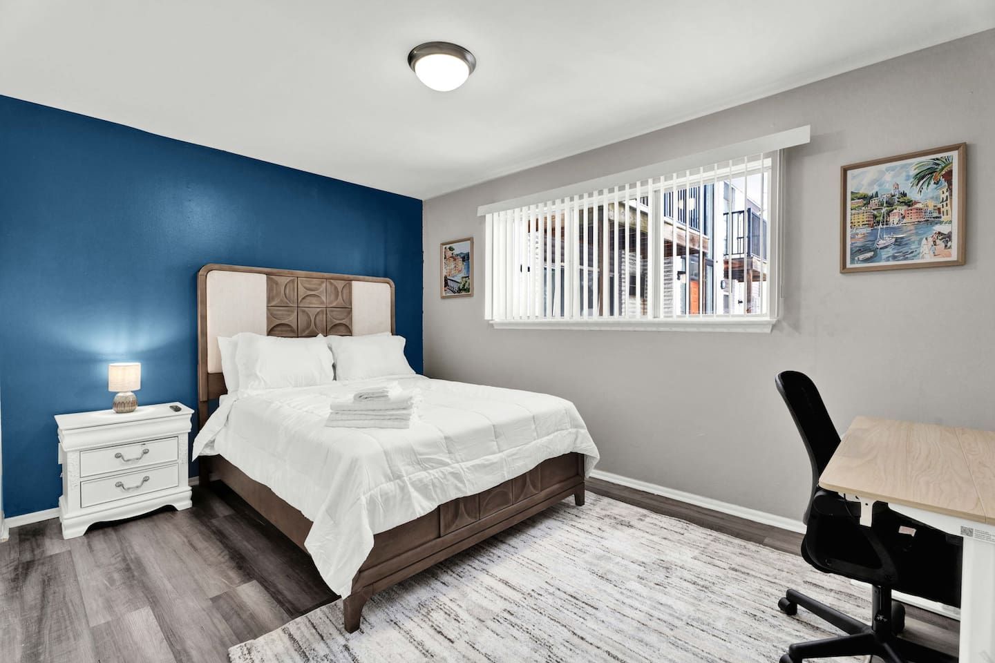 A bedroom with a bed, nightstand, desk, chair, window with blinds, and artwork on the wall.