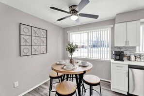 A modern kitchen with a dining area, white cabinetry, a ceiling fan, and a window with blinds. - Sand & Sky Suite | ~Smart TV's ~Full Kitchen (Sterling Heights)