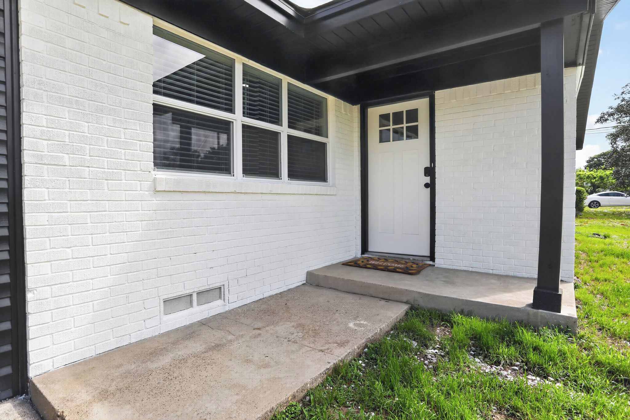 Una casa de ladrillo blanco con puerta y ventanas negras, un porche de concreto y un felpudo.
