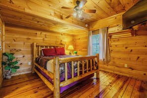 Un dormitorio de cabaña de madera con cama, ventilador de techo y ventana con cortinas.