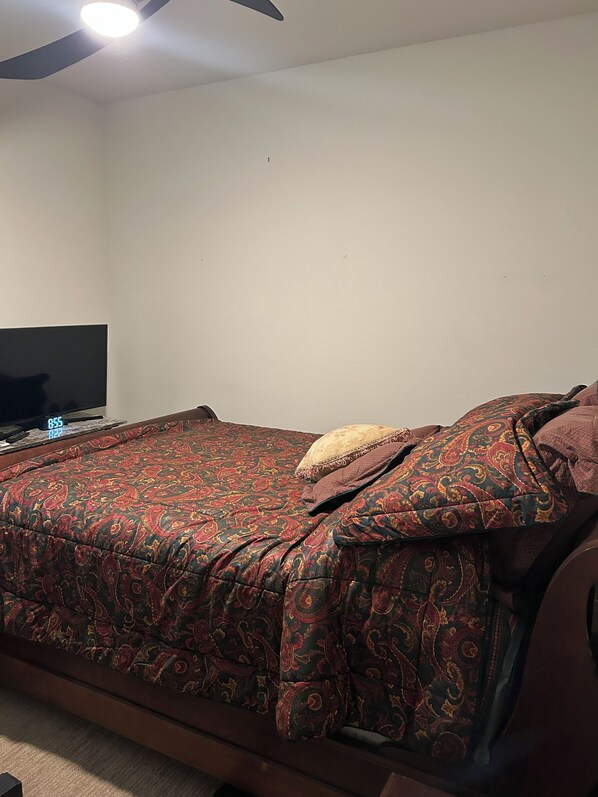 Uma cama com colcha estampada, uma televisão sobre suporte e um ventilador de teto.