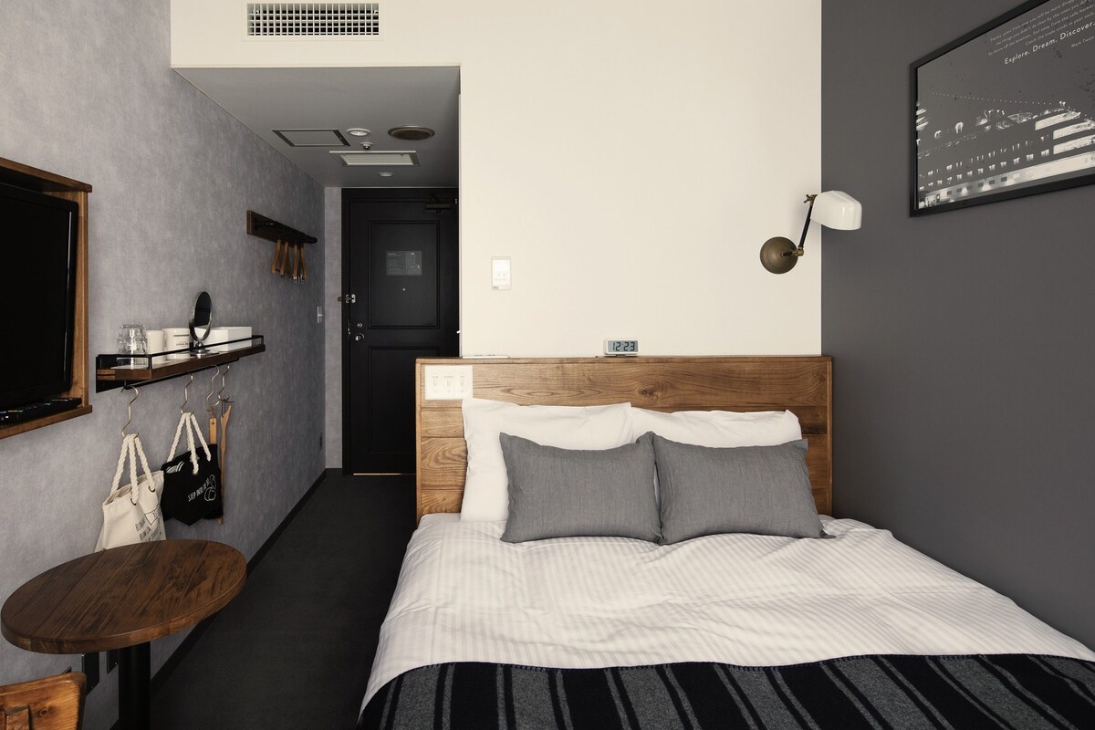 A hotel room with a bed, a small table, a chair, a shelf with bags, a wall clock, and a framed picture.