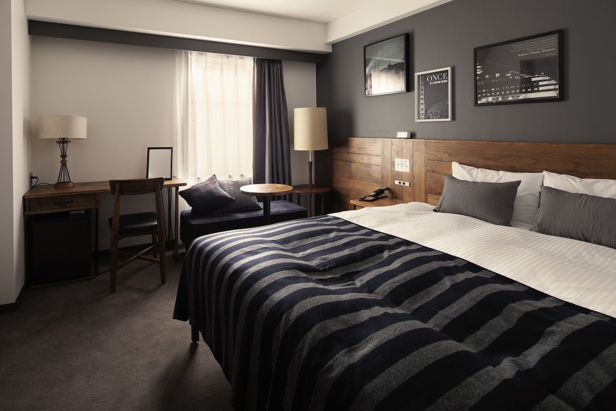 A hotel room with a bed, a desk, a chair, a lamp, and a nightstand.