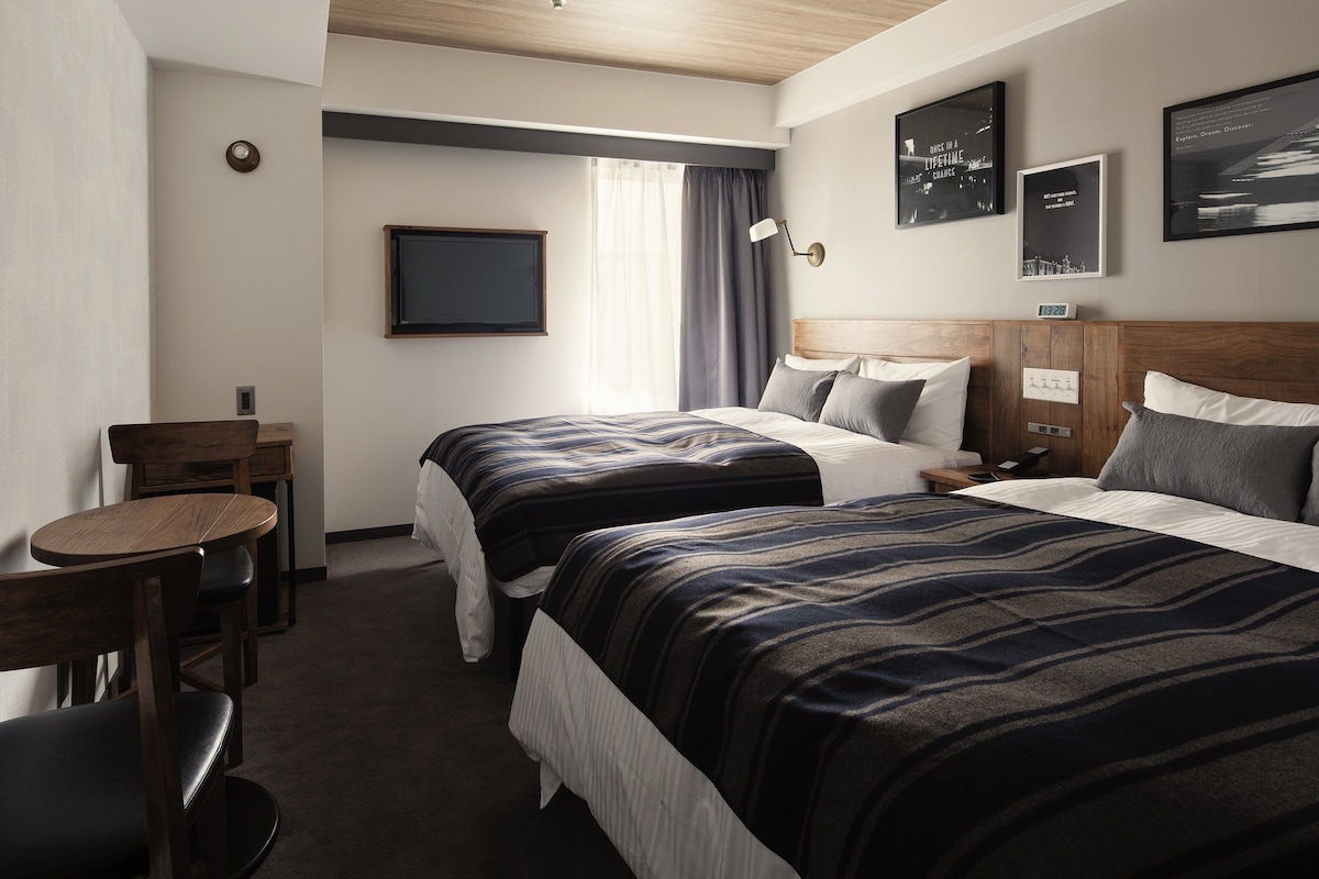A hotel room with two beds, a desk, a chair, and a TV.