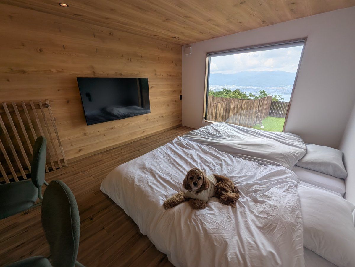 A bedroom with a large window, a bed, a chair, and a television mounted on the wall.
